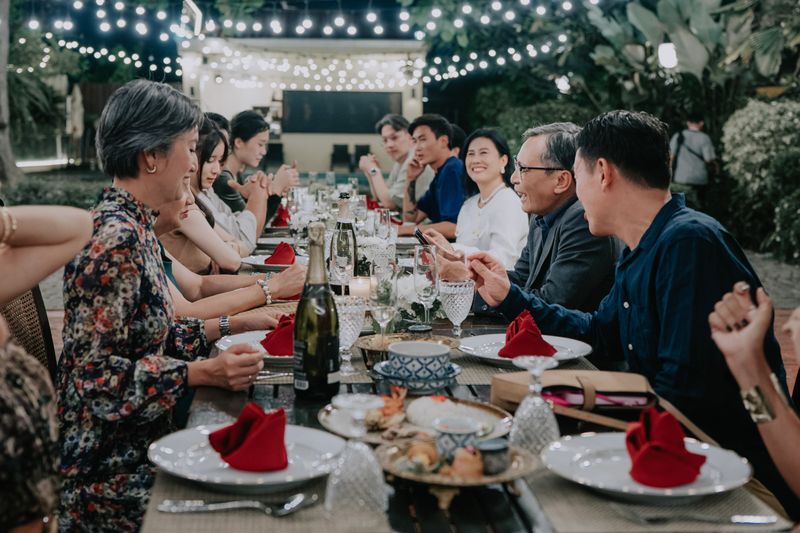 group of multi-generational Asian people sharing a meal and laughing at a long decorated table during an evening garden party. The scene features warm candlelight, wine, and festive string lights in the background.
