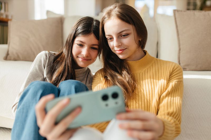 Two girls sit together on a couch and share a smartphone, smiling in soft daylight. Lifestyle concept of friendship, family time, and modern childhood technology at home.
