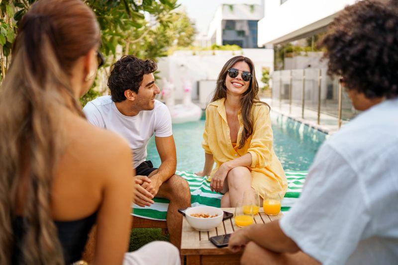 Four diverse friends sitting around a private swimming pool, sharing snacks from a tray. Woman in yellow outfit and sunglasses smiling while conversing. Modern residential backyard setting with white architecture and greenery. Bright sunny afternoon creating a relaxed social atmosphere.