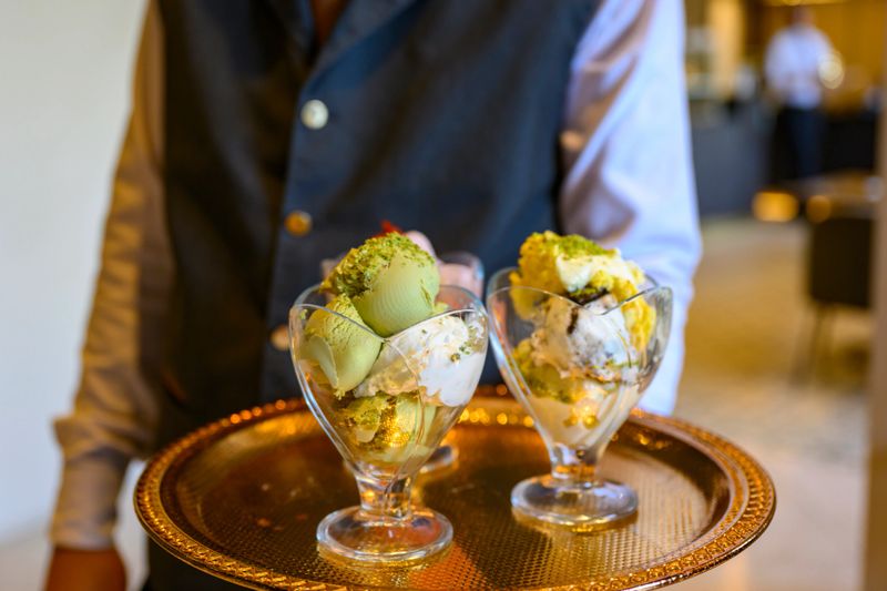 Waiter holding a golden tray with two glass sundaes filled with scoops of pistachio and vanilla ice cream, topped with chopped pistachios, providing an elegant dessert service