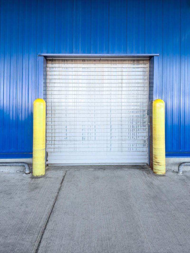 A closed industrial warehouse loading gate is set into a blue metal wall, flanked by two bright yellow safety bollards and a concrete driveway in front. The clean geometric scene is ideal for logistics themes, storage facilities, distribution centers, and industrial safety concepts. Horizontal composition with copy space suitable for web banners, brochures, and editorial layouts.