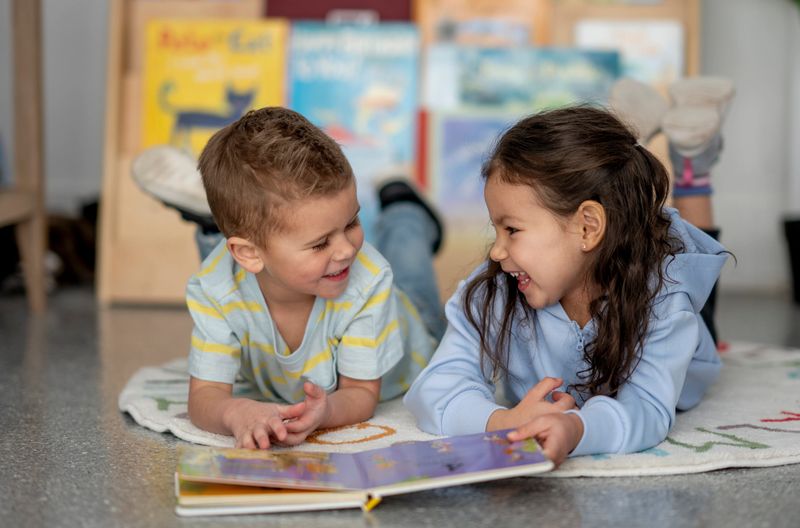 Two kids share a joyful moment as they read a colorful book together on a rug. Their smiles and close collaboration create a warm, playful learning scene.