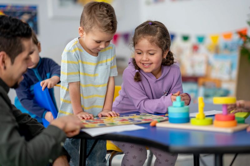 Two young children, a boy and a girl, engage with a colorful puzzle at a table while a male caregiver guides their activity. Bright toys and banners create a cheerful learning environment.