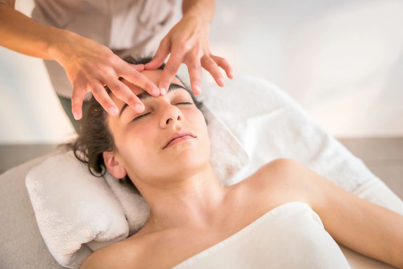 Young adult woman closing eyes and relaxing on a spa bed while a therapist performs a facial massage, providing wellness, therapy, and skincare for beauty and stress relief