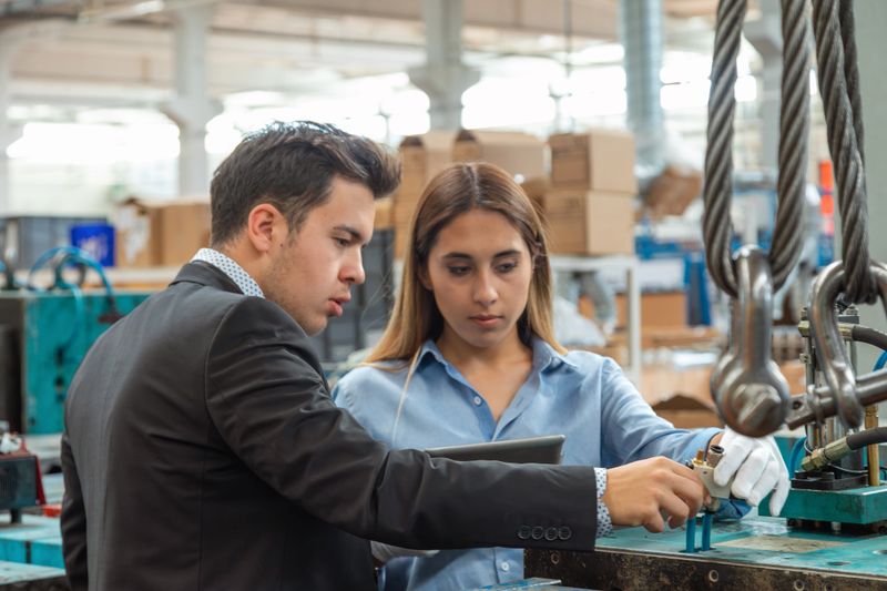 Focused teamwork scene showing professional engineer and business supervisor inspecting heavy industrial equipment and discussing technical details on digital tablet inside manufacturing plant, collaboration between management and engineering roles with realistic workplace environment and copy space