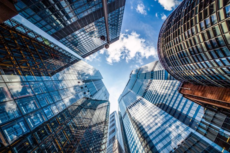 Dramatic low angle view of modern glass skyscrapers surrounding the viewer in a dense urban business district. Reflective facades and curved architectural lines create a powerful perspective toward the blue sky, symbolizing corporate growth, innovation, finance, and future city development. Ideal for business concepts, technology, investment, real estate, and abstract urban backgrounds.