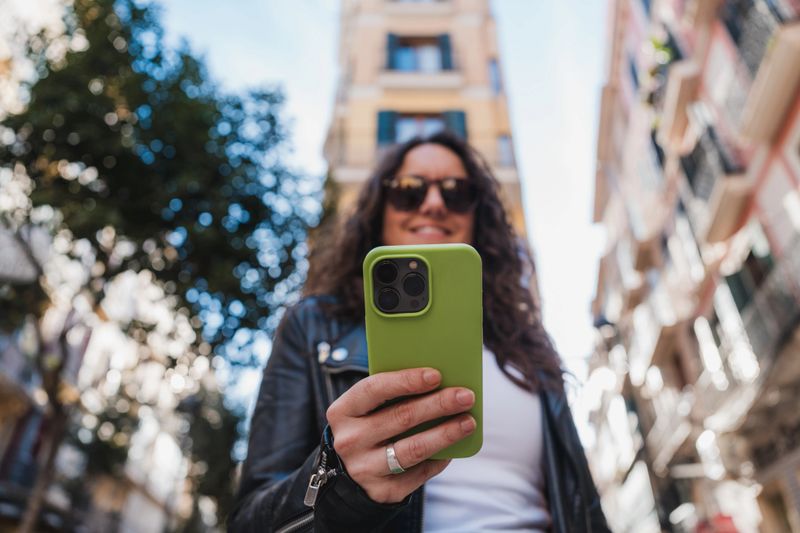 Woman smiling, holding smartphone with green case, wearing sunglasses and leather jacket
