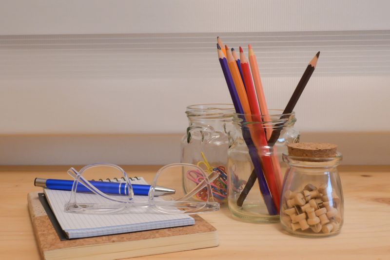 Organized desk with notepad, reading glasses, pen and colorful pencils with paper clips and pushpins on cork and wood surface, warm natural light suggests study and creativity