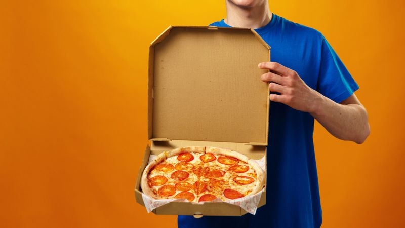 A man stands holding a pizza box filled with pepperoni pizza in front of an orange wall.