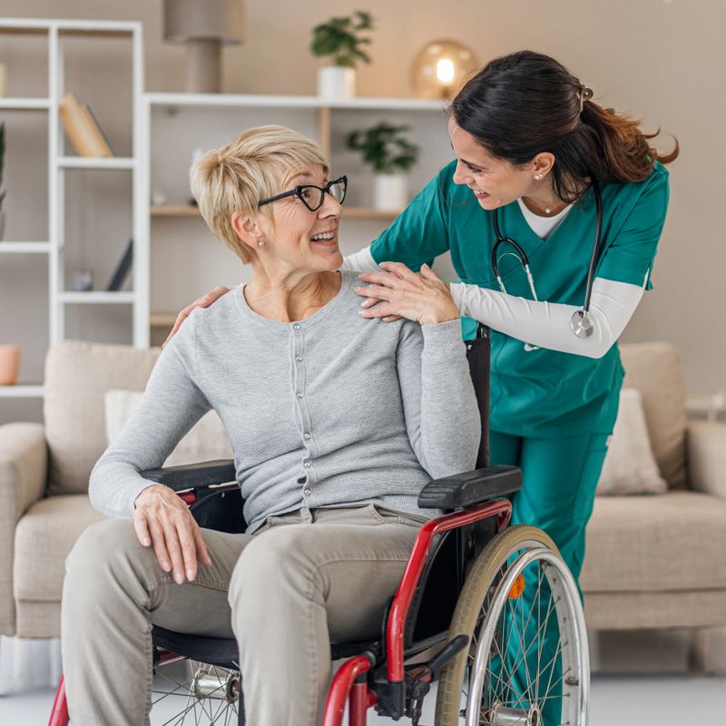 Caring nurse supporting a happy mature adult woman in a wheelchair at home, providing professional assistance and comfort