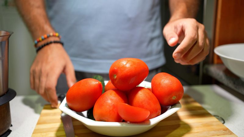 Hands arranging ripe tomatoes in bowl on wooden board beside ceramic dish, vibrant red skins glistening, wrist bracelets visible, kitchen prep for dinner, farm
