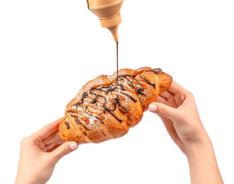 Sweet croissant topped with caramel and nuts while chocolate syrup is poured over it, held by woman hands on white background.