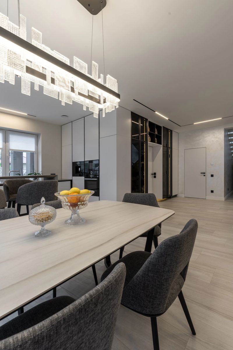 Elegant vertical shot of a modern open-plan dining room. Features a marble-top table, grey chairs, and a striking crystal chandelier. The background showcases a sleek kitchen, linear ceiling lighting, and a minimalist white hallway with a door.
