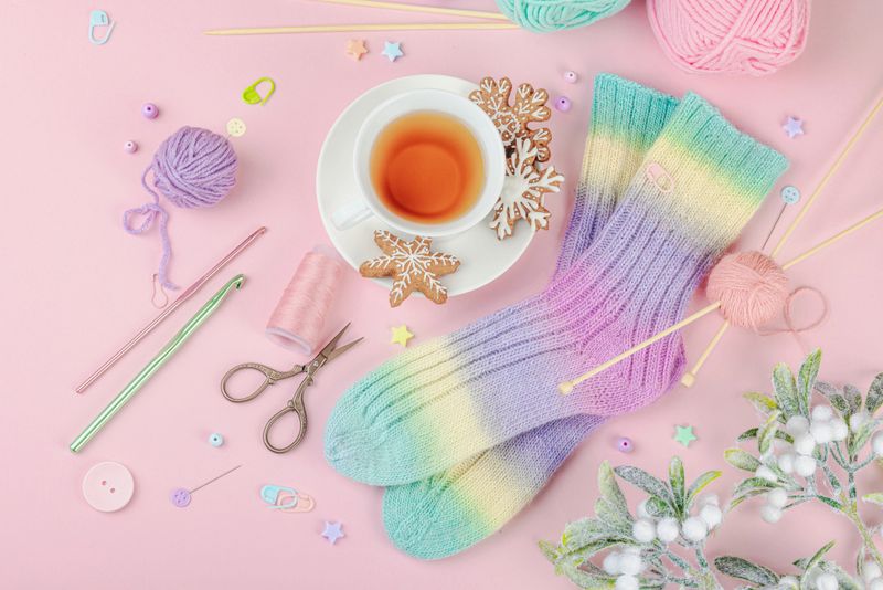 Handmade knitted socks with rainbow gradient on pink background. Cozy composition with cup of tea, gingerbread cookies, yarn balls, crochet hooks and knitting needles, top view