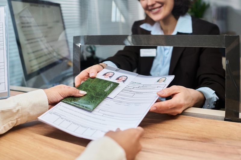 Caucasian woman sitting behind counter assisting customer by receiving passport and visa application documents at visa center, hands of young adult visible handing over paperwork