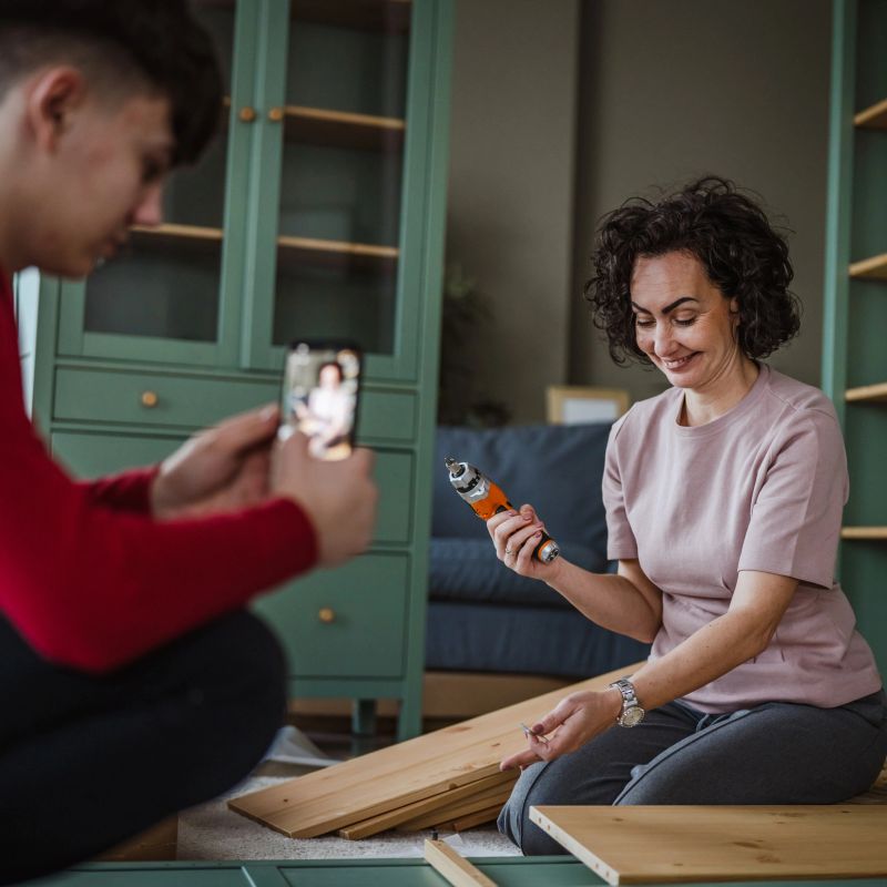 Son capturing video of his smiling mother constructing new wooden shelves with an electric screwdriver, sharing their diy home project and bonding experience