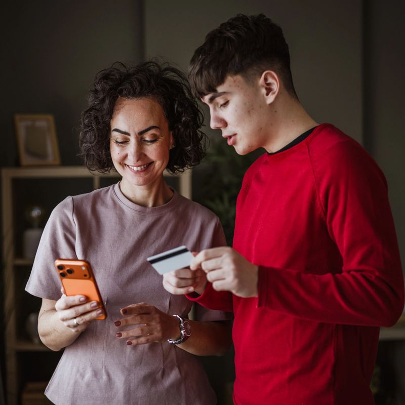 Smiling mother and son standing at home, sharing a smart phone and credit card while making a secure online purchase, enjoying convenient digital shopping and family connection