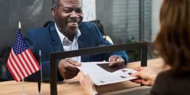 Man smiling while handing over documents through a glass window.