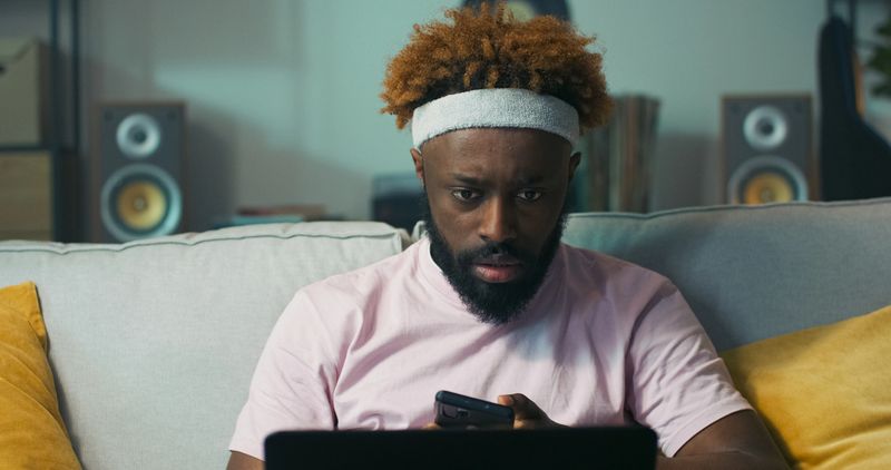 Bearded guy with warm skin tone and curly hair sits on the couch with phone and laptop. He reads a notification confirming he got the job and smiles with joy.