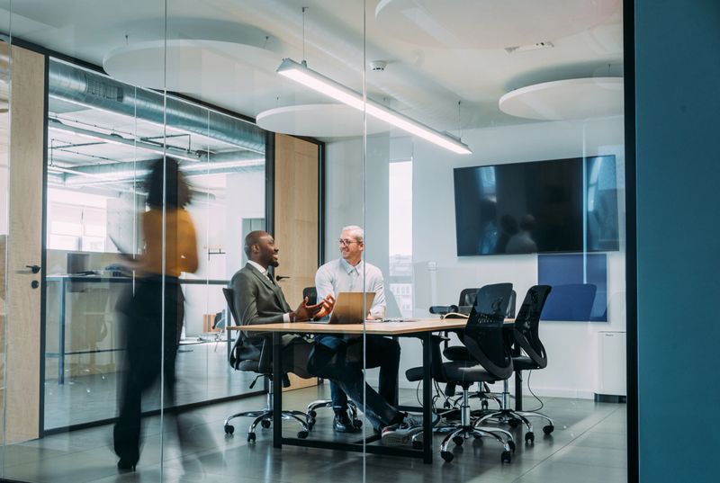 Shot of business people having a meeting in modern office. Blurred motion of energetic businesspeople on the move and project team members discussing ideas in the office board room. Businesspeople walking in blurred motion around their colleagues. Business persons having a business meeting in board room while their colleagues are walking in blurred motion. Creative business team on meeting in the office. The view is through glass.