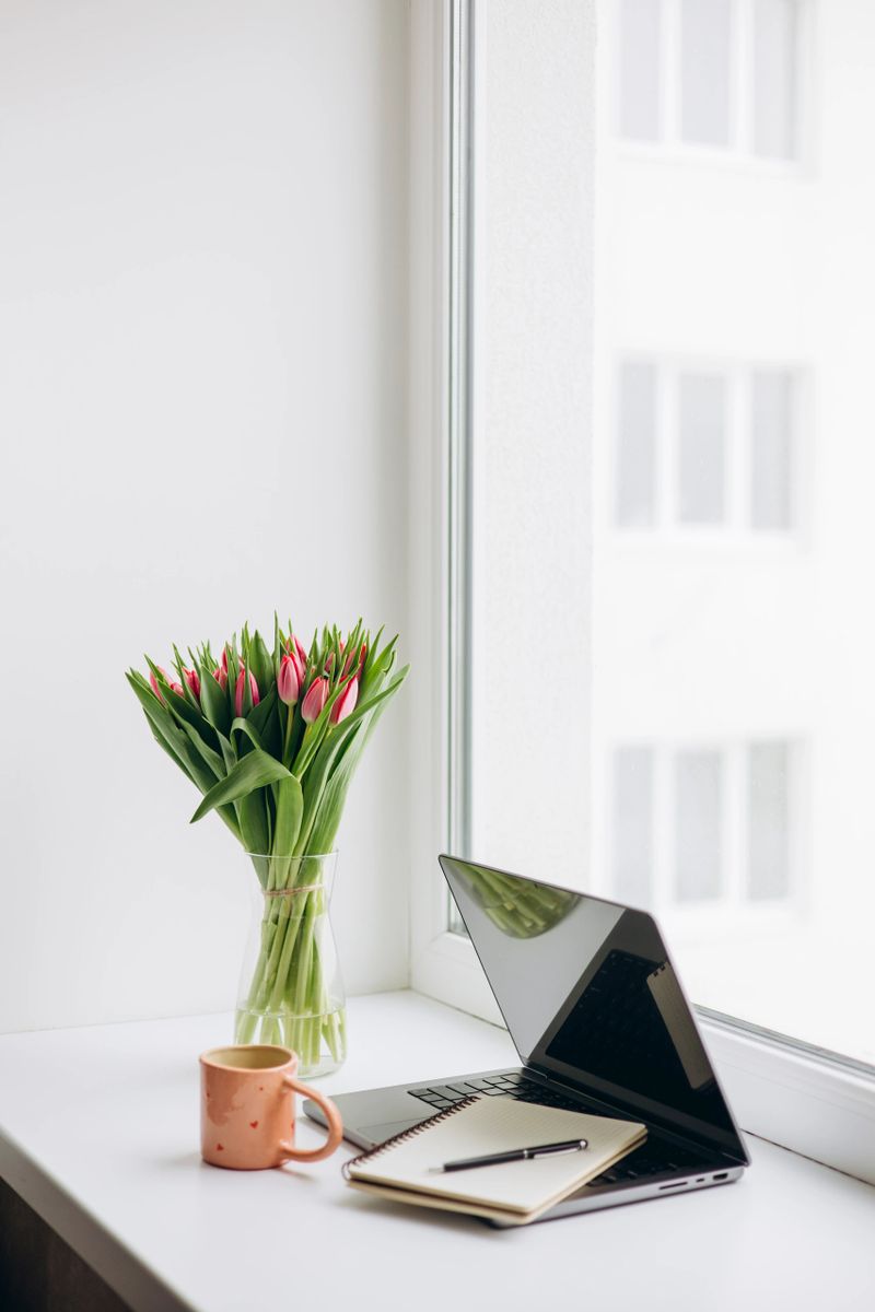 Minimal home office desk with laptop, notebook and coffee near window. Fresh tulips in vase, natural light and calm remote work lifestyle concept with copy space.