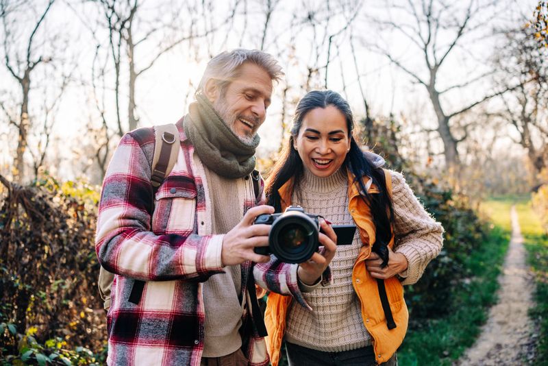 Two people explore the outdoors while hiking a sunny trail. They share smiles as they look at photos on a camera. The fresh air and trees surround them.