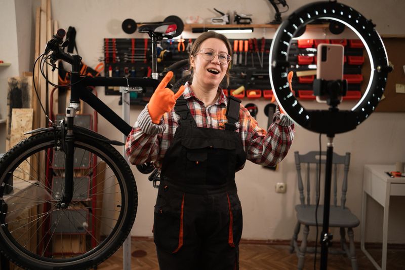 Bike mechanic in garage. Female worker blogger reviewing and recording video on smartphone with rock hand gesture for wild expression, devil horn sign, funky emoji in bicycle workshop. copy space
