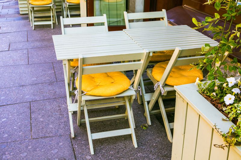 Inviting cafe setup with light wooden tables and chairs, accented by vibrant yellow cushions. The scene exudes warmth and relaxation, perfect for a leisurely coffee break or casual meeting.