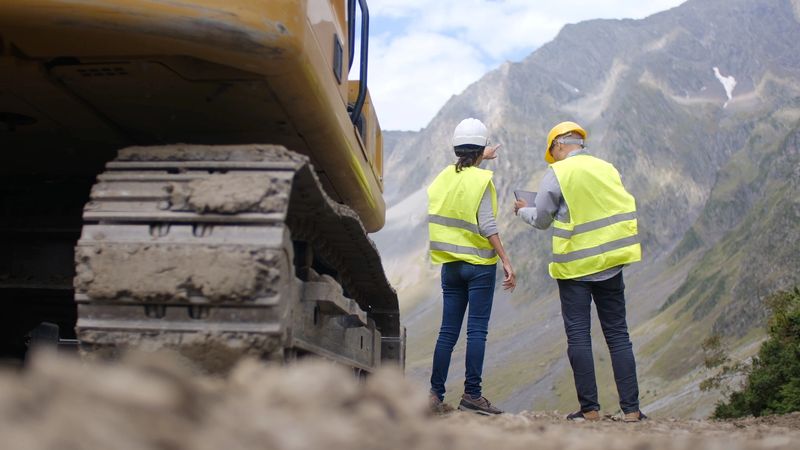 Two workers wearing hard hats and safety vests standing at a construction site in the mountains, discussing project plans with a digital tablet and pointing towards the landscape