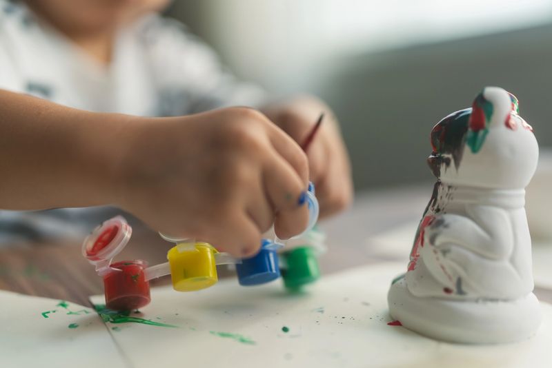 Close-up of an Asian toddler’s small hands painting a ceramic figurine with colorful paints at a table indoors. The child uses red, yellow, blue, and green paint pots to create a fun art project, showing creativity, fine motor skills, and early childhood development. This candid home activity highlights learning through play, artistic expression, preschool education, and nurturing parenting in a bright, cozy family environment.