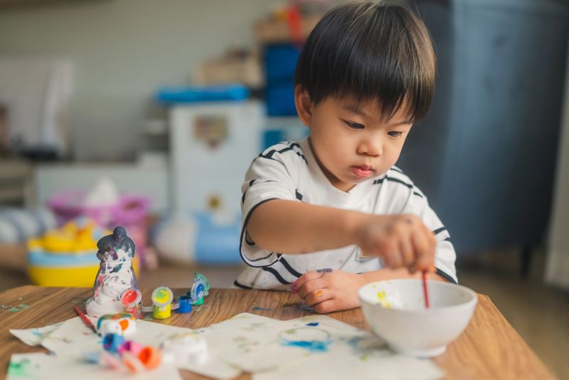 Asian toddler boy painting at home table, focused on creative art and craft activity, developing fine motor skills and early childhood education through colorful DIY ceramic project in bright natural light family lifestyle setting.