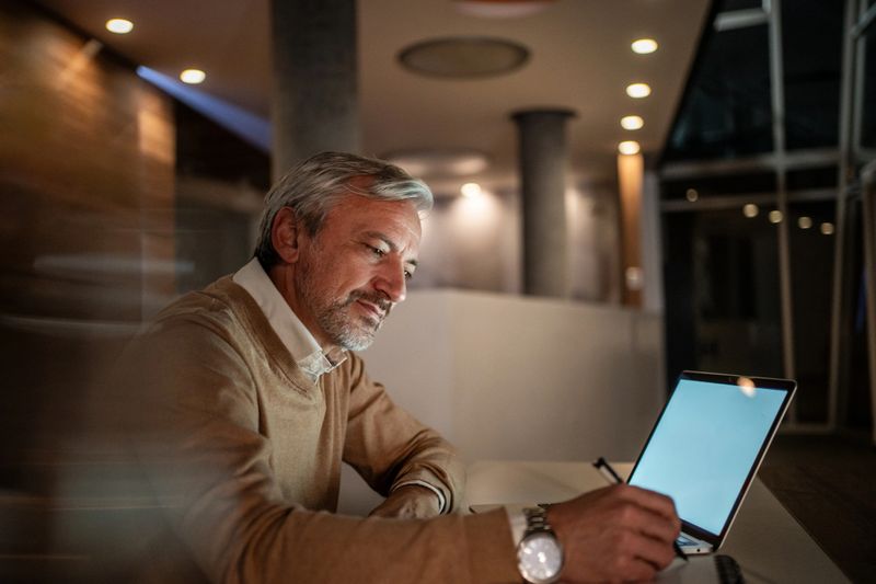 Mature man working late on a laptop at a dim modern office desk, focused and writing with a stylus, warm desk light illuminating his concentrated, professional expression