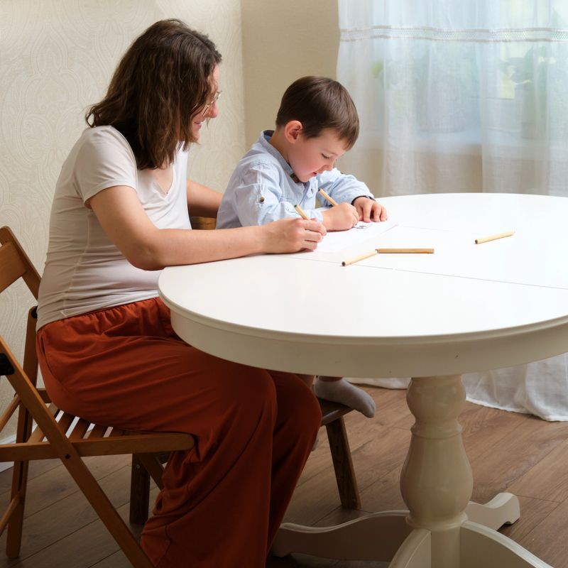 Woman and a small boy cheerfully coloring a picture on the white table. Cute little child engaged in creative activity with his mother at home. A child aged 4 years (four years old)
