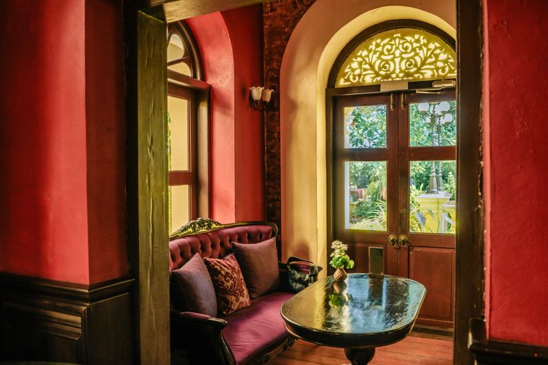 vintage colonial style lounge interior featuring red walls, a velvet sofa, and an arched wooden door with decorative glass, creating a refined boutique hospitality atmosphere, Bangkok