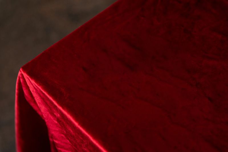 Close-up detail of a deep red velvet tablecloth draped over the corner of a table, showing soft fabric texture and gentle folds against a blurred neutral background.