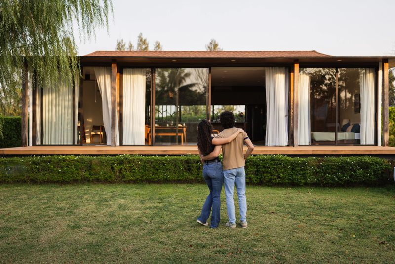 Rear view of a young couple standing together in their backyard with arms around each other, admiring their newly purchased modern home.