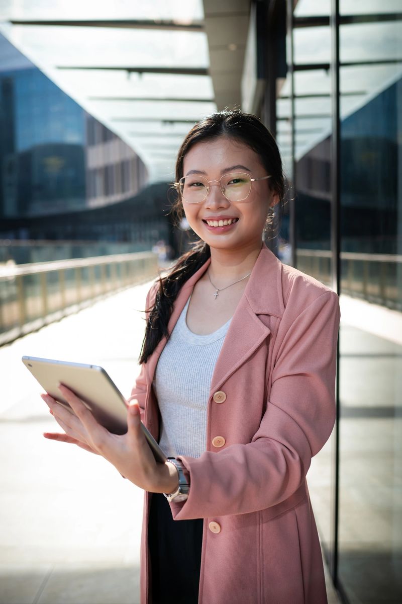A young Asian businesswoman with glasses smiles while holding a tablet outdoors amidst the towering structures of the financial district.