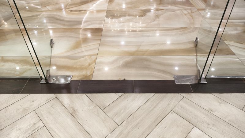 A high-angle view of a modern interior floor. Glossy beige marble-effect tiles meet matte wood-look tiles in a herringbone pattern. A frameless glass door with metal fittings separates the two areas.