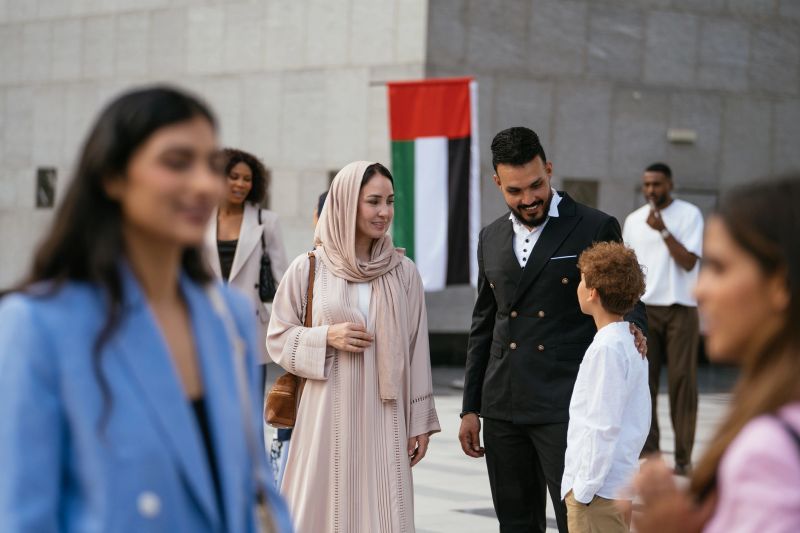 A family interacts outdoors in an urban area, with a woman in traditional attire, a man, and a child, set against the backdrop of the UAE flag.