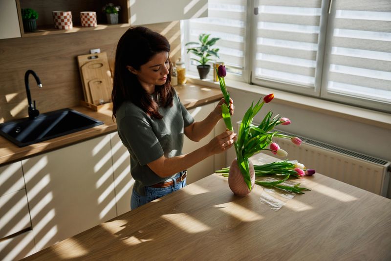 A Caucasian woman lovingly arranges a colorful tulip bouquet on a sunlit kitchen island, creating a simple spring flower decoration at home to brighten the space and relax after a busy day.