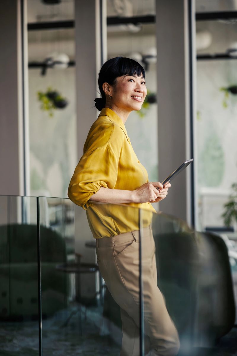 Mid adult asian businesswoman smiling confidently and looking away while holding a digital tablet, standing in a contemporary office space, symbolizing future vision and success
