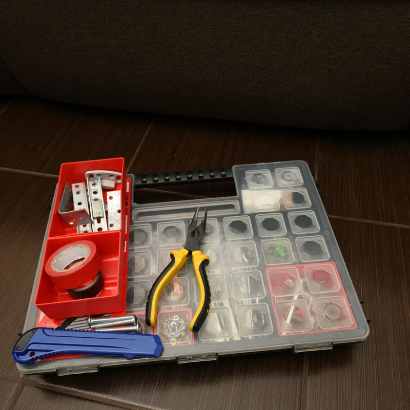 A storage box holds various tools and supplies on a wooden floor beside a sofa. Items include tape, pliers, and fasteners.