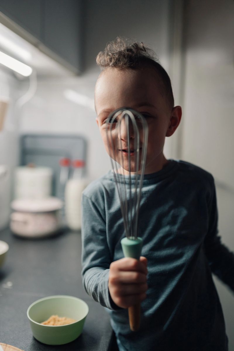Young boy joyfully pretending to be a pirate with a whisk covering one eye, flashing a big smile in a warm and inviting kitchen setting, embracing the joys of baking and discovering new things