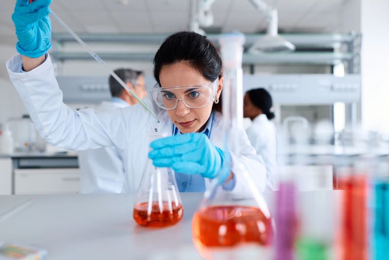 Female scientist wearing safety goggles and gloves using a pipette to carefully transfer orange liquid into an Erlenmeyer flask during a chemistry experiment in a modern research laboratory