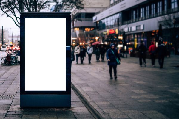 Empty vertical billboard on a city sidewalk with pedestrians in the background.