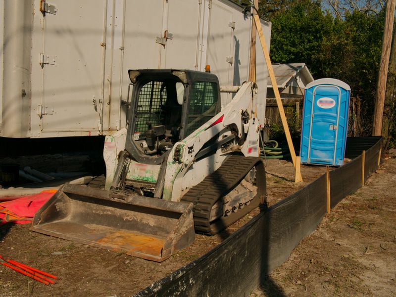 A compact skid steer loader sits next to a white work trailer and blue portable toilet behind a silt fence on an outdoor construction site, highlighting equipment, temporary facilities, and groundwork.