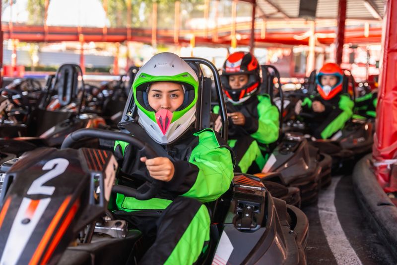 Group of women in racing suits and helmets seated in go-karts on the starting grid, focused and smiling as they prepare for a competitive, fun-filled motorsport race