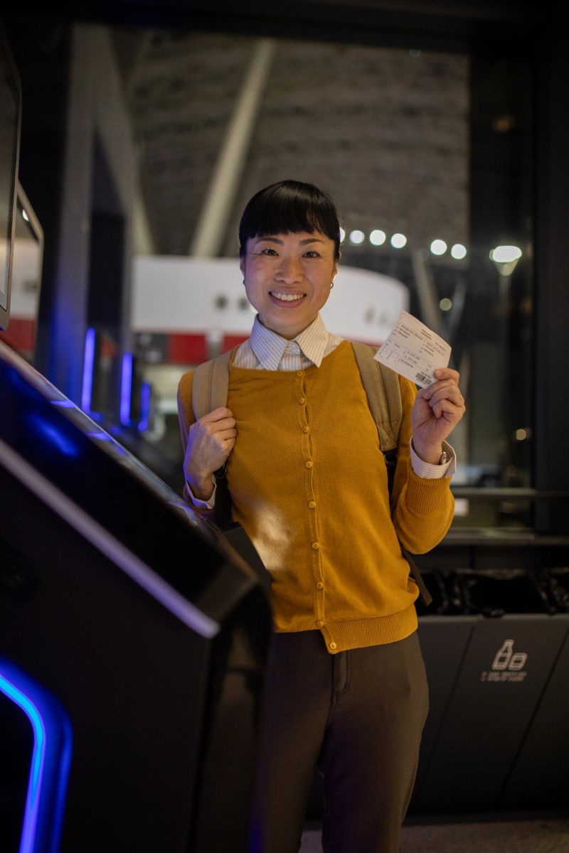 Happy young woman smiling at camera while using a self check-in kiosk at the airport, holding her boarding pass and ready for travel, confident and casual before departure