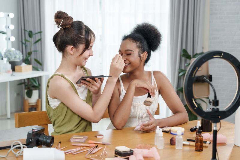 Teen beauty bloggers in diverse friendship smiling while reviewing makeup palette and skincare items during home live streaming session in bright room with camera and ring light