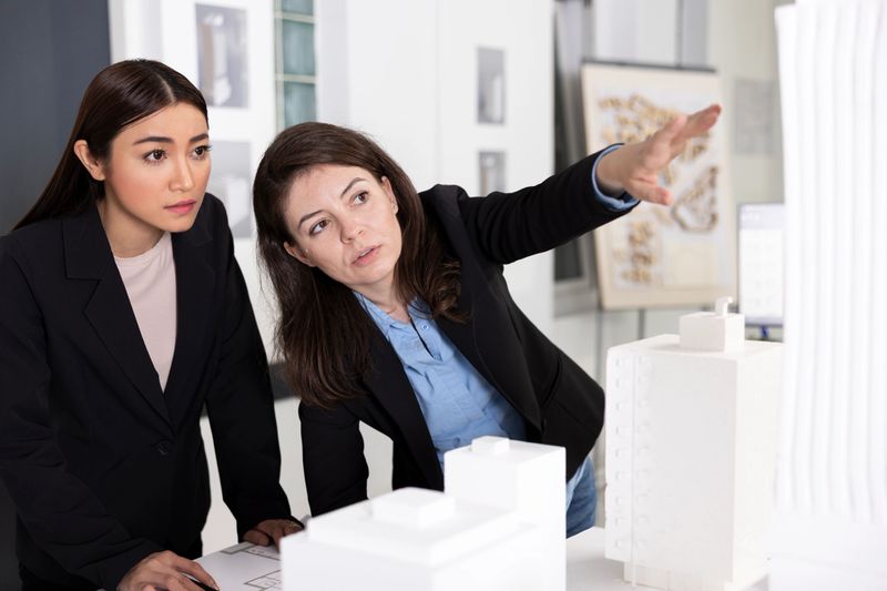 Architects analyze detailed architectural model while discussing structural ideas and project layout. Professional women show teamwork, precision and creative problem solving in modern design office.